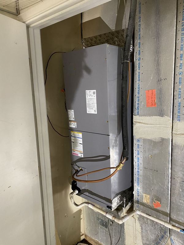 Gray air handler installed in a utility closet next to new foil-faced duct board, with insulated refrigerant line and PVC condensate drain