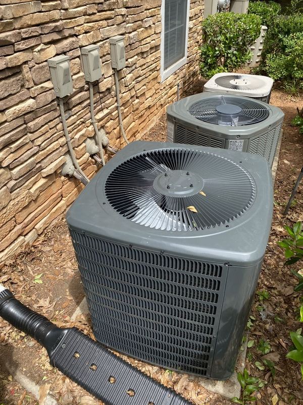 Three outdoor AC condensers lined up beside a stone-veneer wall with disconnect boxes
